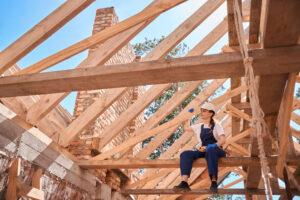 A multifamily general contractor sitting on beam of roof frame and looking at construction site.